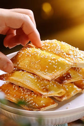 Hand picking up a piece of golden-brown fried pastry with sesame seeds on a white plate.