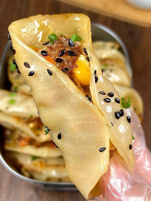 Handheld dumpling with fillings and sesame seeds, with a bowl of more dumplings in the background.