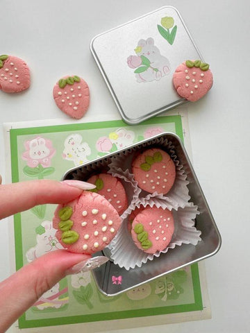 Pink strawberry-shaped cookies in a tin with decorative elements on a white background