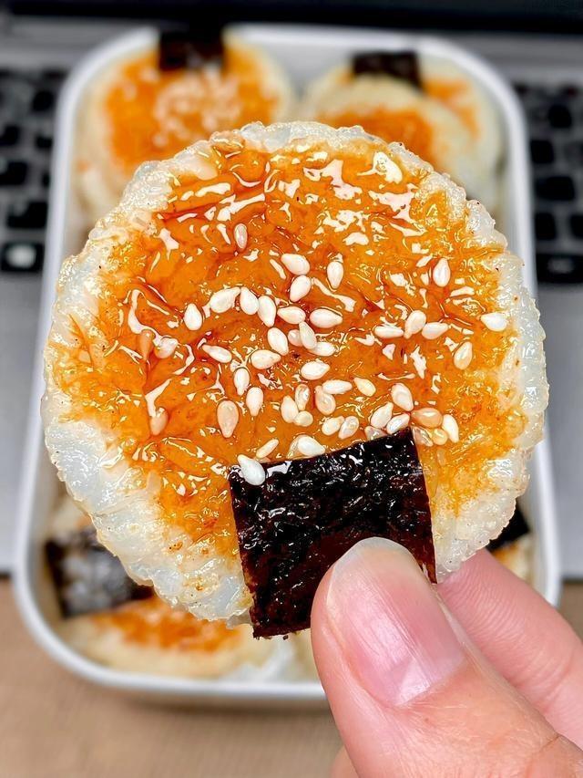 Hand holding a piece of rice cake with orange topping and sesame seeds, with more rice cakes in the background.