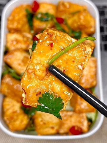 Fried tofu with garnishes being held by chopsticks over a container of more tofu.