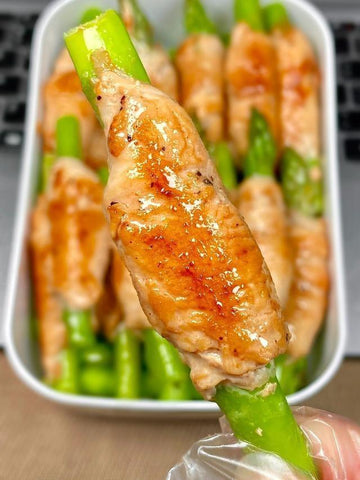 Chicken wing with asparagus on a toothpick held by a hand, with more wings in the background.