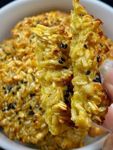 Close-up of a hand holding a piece of crispy rice cake with sesame seeds, with more rice cakes in the background.