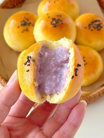 Hand holding a half-eaten purple yam bun with other buns in the background.