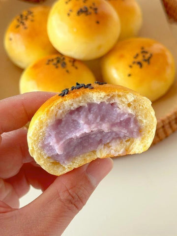 Hand holding a half-eaten purple yam bun with more buns in the background.